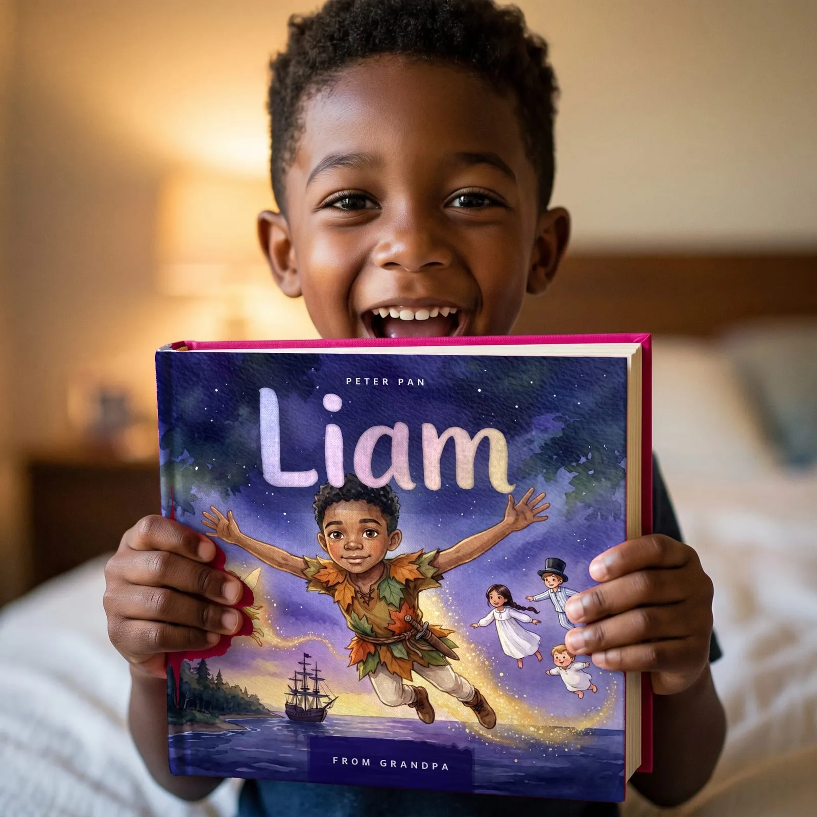 A boy holding up his hardcover Peter Pan book, delighted. The cover shows him flying over Neverland with Tinker Bell, and the name Liam is painted across the night sky.